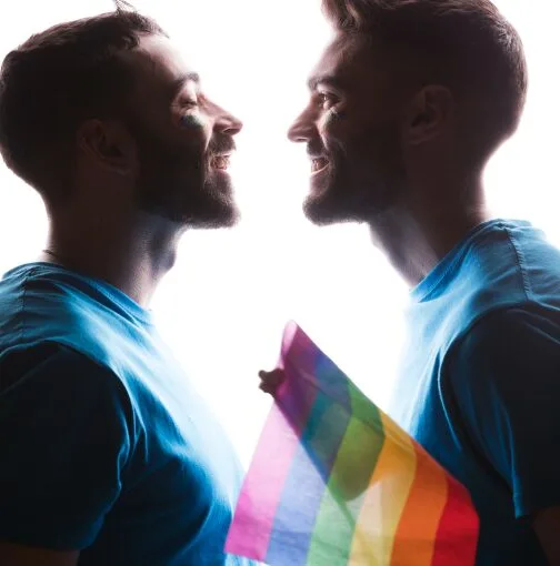 Gay couple lovingly gazing into each other’s eyes, holding pride flag, promoting therapy for gay men