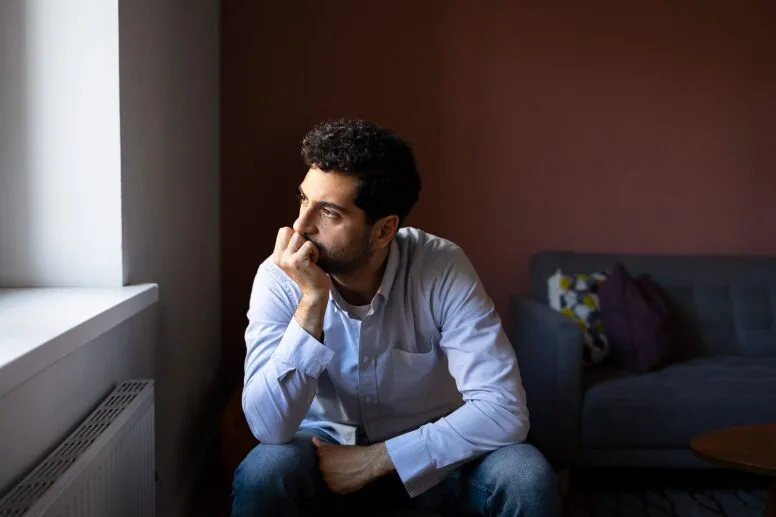 Man sitting in a dark room looking outside the window pondering on taking online therapy in Colorado