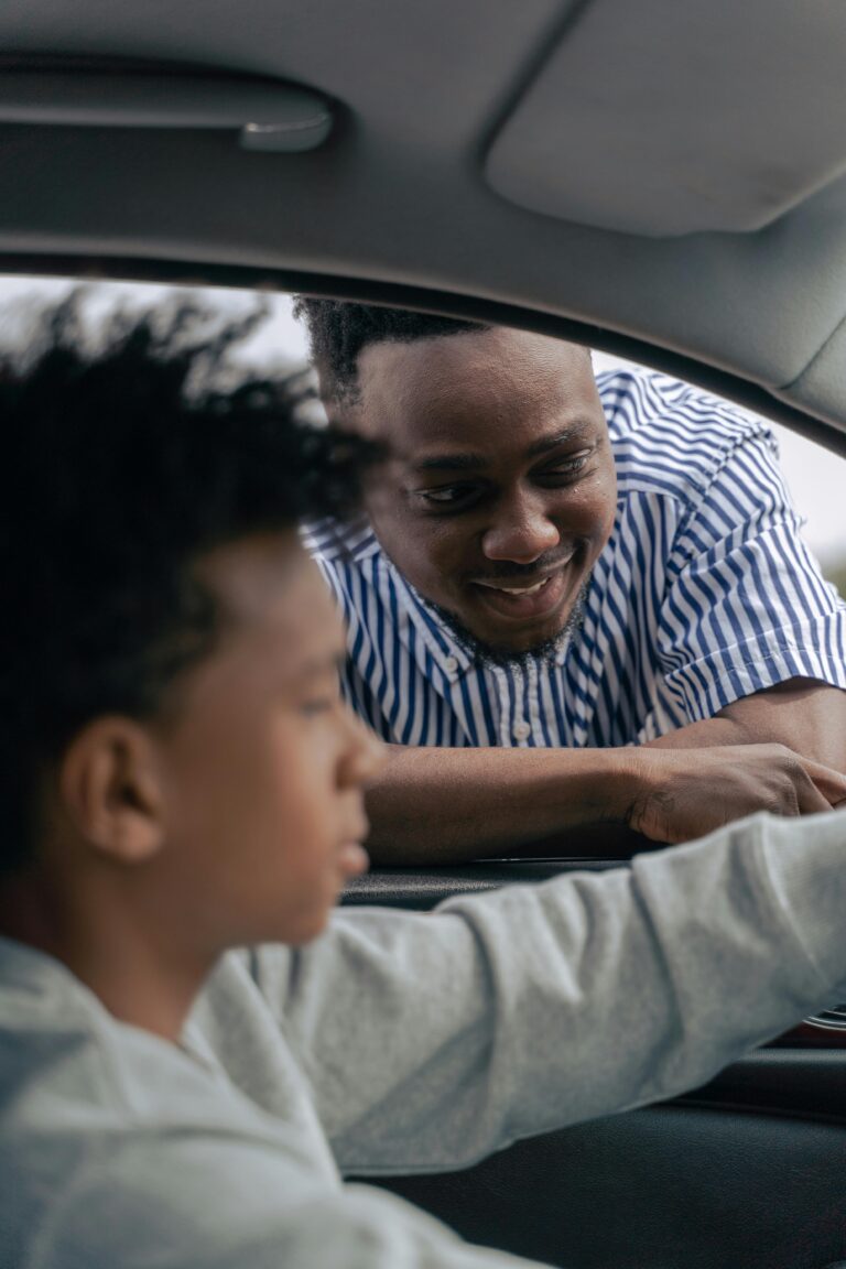 Father leans into a car window, smiling and coaching his teen son in the driver’s seat during a supportive driving lesson